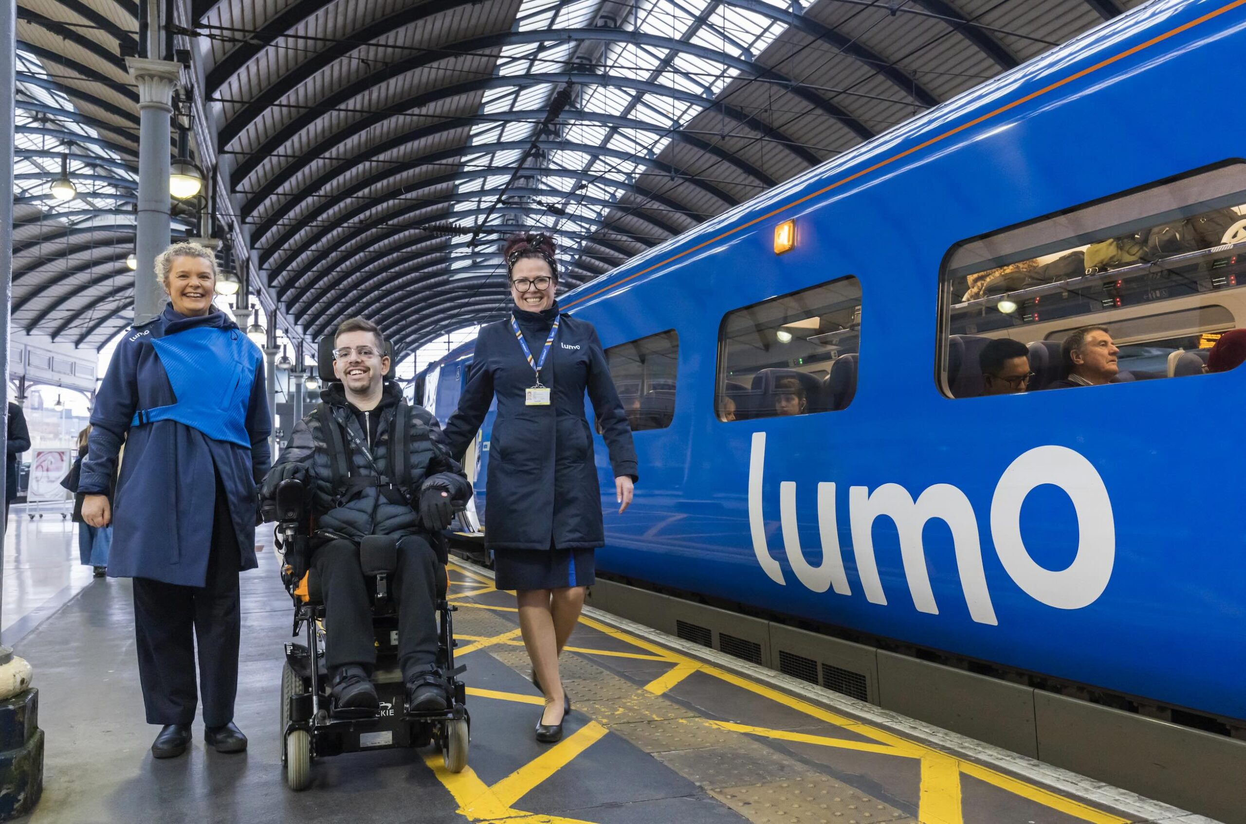 Three people, one in a wheelchair alongside a Lumo train