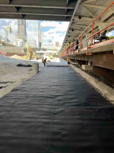 Workers unrolling a black geocomposite membrane across a gravel trackbed beside an elevated rail platform, with excavation equipment and the Chicago skyline in the distance.