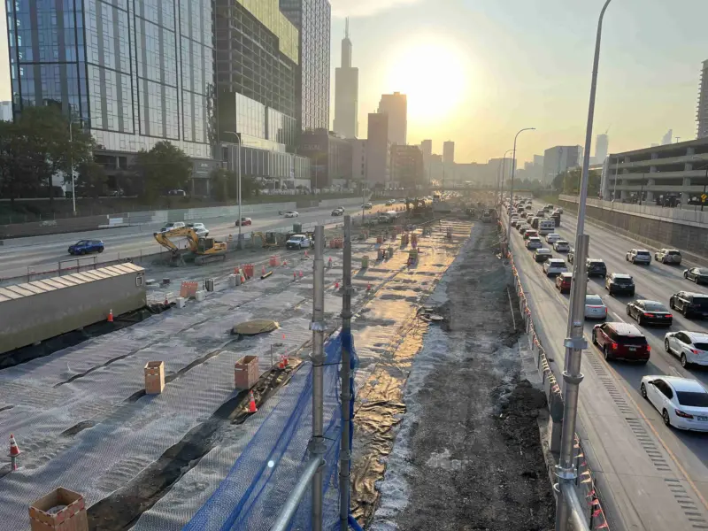 Construction workers in high-visibility clothing rolling out a wide black TrackTex geotextile membrane along a railway trackbed at UIC-Halsted Station, with the Chicago skyline and an excavator in the background.