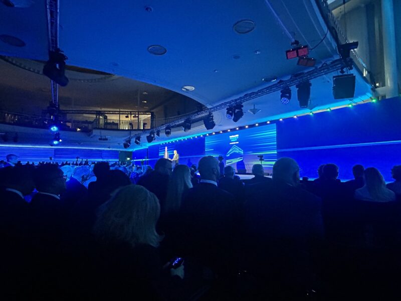 Wide shot of the InnoTrans opening ceremony hall, with two figures shaking hands on stage in front of a screen reading "The Future of Mobility."