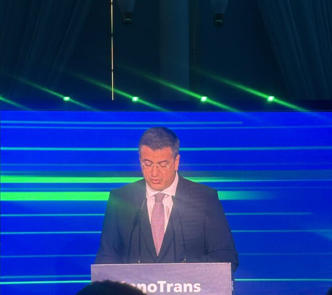 Apostolos Tzitzikostas delivers remarks from the InnoTrans podium, lit against a blue-and-green backdrop.