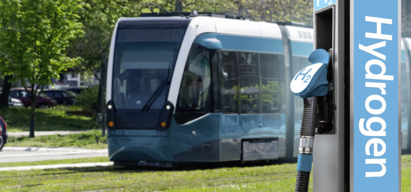A blue and white tram runs on a grass-covered track past leafy trees, with a hydrogen refueling pump in the foreground. The pump nozzle is marked "H₂" and the pump column carries a vertical "Hydrogen" label.