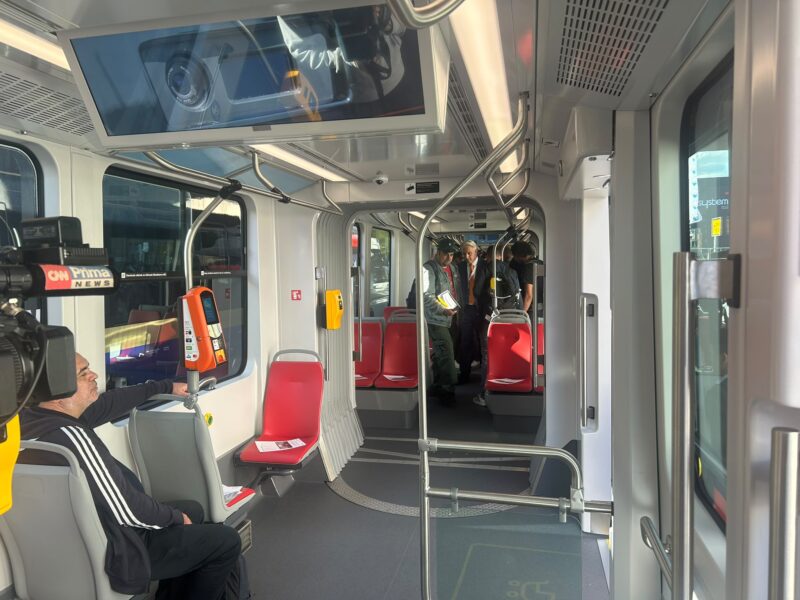Inside the low-floor tram, showing the spacious, accessible interior with red seating, grab rails and onboard passenger information screens as journalists and guests tour the vehicle.