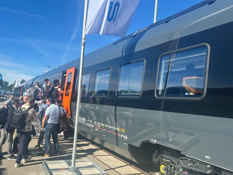 Trade fair visitors queue to board the FLIRT Nordic Express at Stadler's outdoor stand at InnoTrans 2026 in Berlin. The train's dark grey livery is accented with bright orange doors.