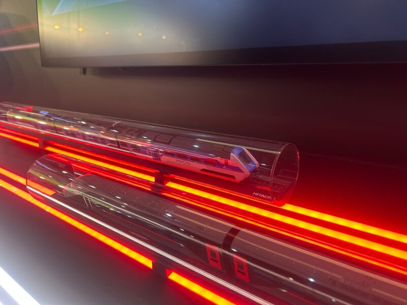 Scale model of a blue and white Intercity battery-electric train inside a clear acrylic tube, lit by red LED strips on the Hitachi Rail stand at InnoTrans 2026