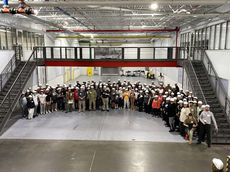 Large group of students and staff wearing white hard hats posing in a factory hall beneath a mezzanine with a "Welcome to Manufacturing Day" banner