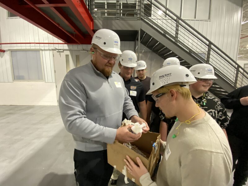 SEKISUI KYDEX team member in a white hard hat and safety glasses examining a student's cardboard egg-drop device as other students look on
