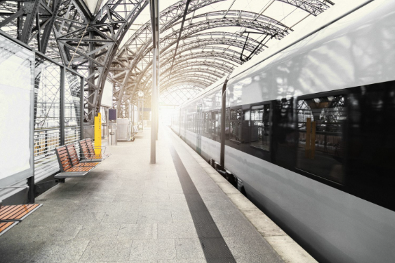 A modern silver passenger train waits at an empty platform beneath a curved steel-and-glass station roof, with wooden benches on the left and bright light at the far end.