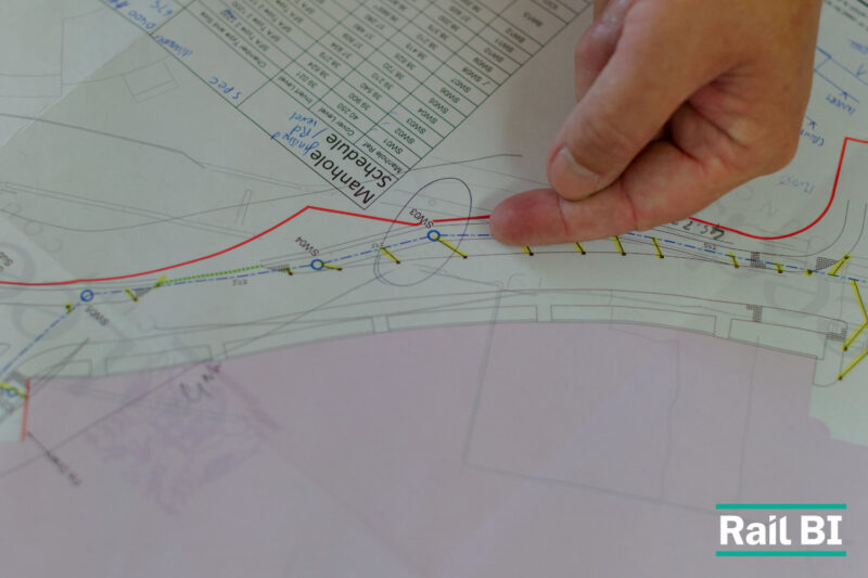 A hand pointing to a marked-up infrastructure planning drawing with a manhole schedule, branded with the Rail BI logo