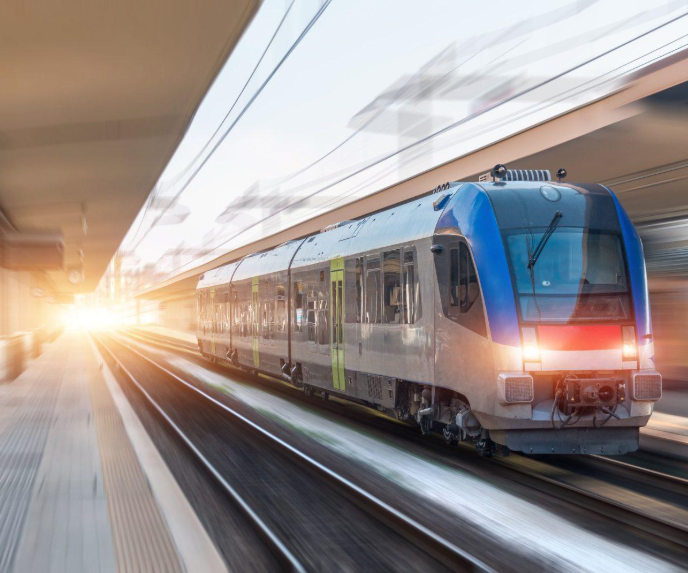 A modern blue, white and silver passenger train speeds through a station platform, with motion blur and bright sunlight flaring at the far end of the platform.