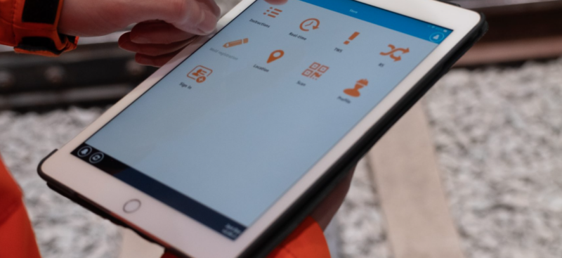 Person in orange high-visibility clothing holding a tablet beside a railway track. The screen shows a rail safety app menu with orange icons, including Instructions, Real-time, TWS, Location, Sign in, Scan and Profile.