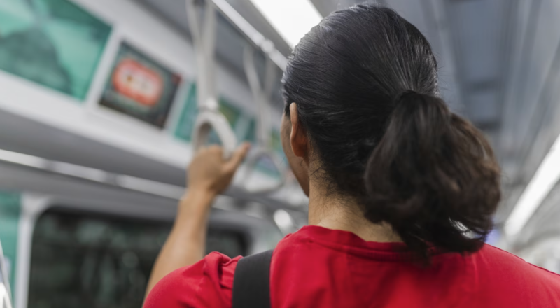 A commuter in a red T-shirt, seen from behind, holds an overhead strap while standing inside a train carriage.