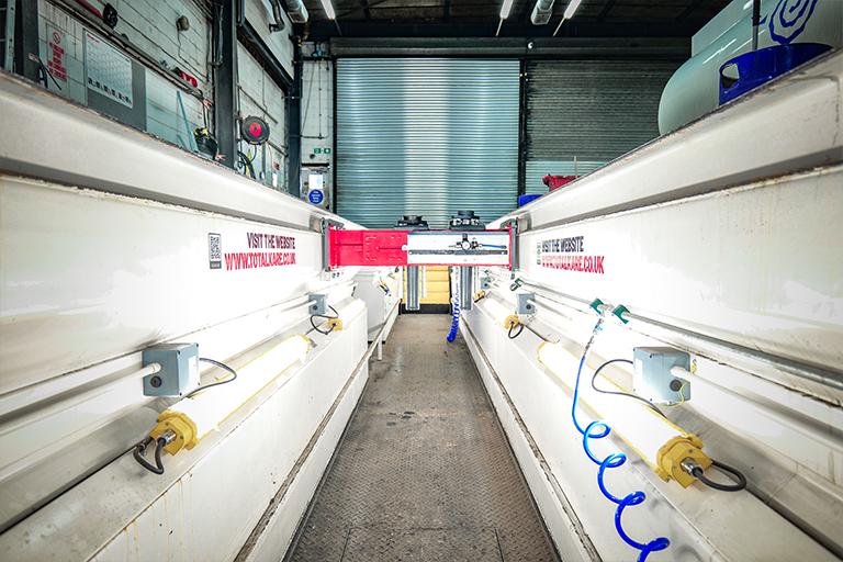 View along a white-walled vehicle inspection pit lit by strip lights on both sides, with a red Totalkare twin-ram pit jack mounted on rails at the far end, blue coiled air hoses and a roller shutter door beyond