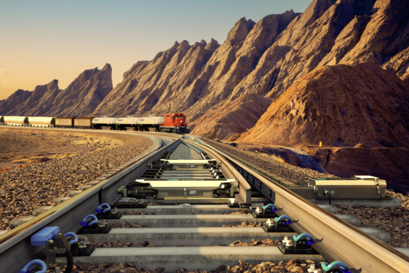 Visualisation of the voestalpine UNISTAR HR switch setting system installed between the rails of a turnout on concrete sleepers, with a red freight locomotive hauling tank and hopper wagons through a rocky desert landscape at sunset