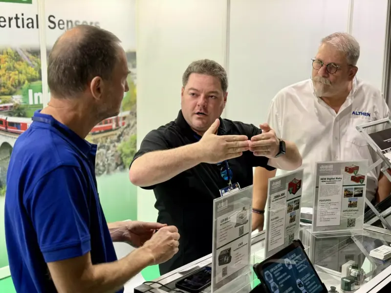 Three men talking at the Jewell Instruments trade show stand. A man in a black polo gestures while explaining something to a visitor in a blue polo, as a man in a white Althen shirt listens. In the foreground, display stands show product sheets for the new Digital Ruby MEMS accelerometers and inclinometers, the JMHA-300 Ruby MEMS accelerometer and the LCF-501-R and LSOX sensors.