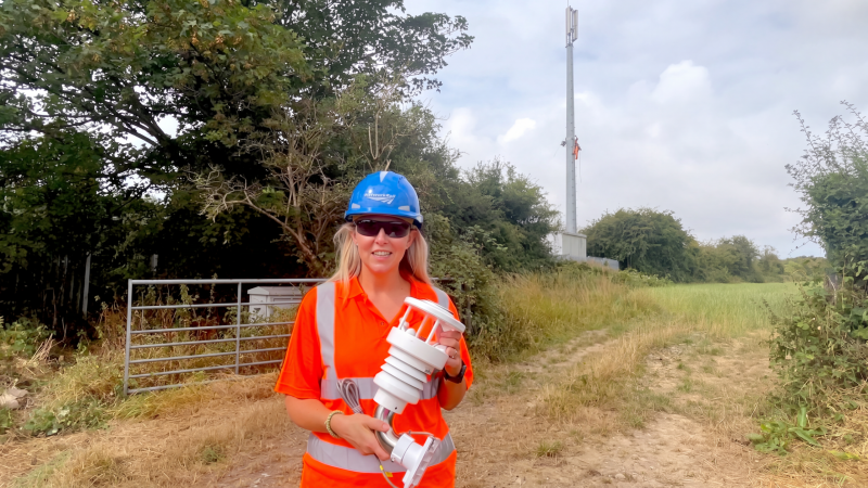 Worker in orange hi-vis and a hard hat holding a 900IoT Weather Station in front of a GSM-R railway communication mast