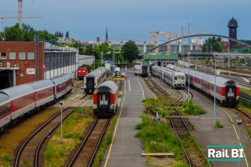 Elevated view of a railway depot with several rows of red and white passenger carriages stabled on parallel tracks, a red shunting locomotive beside a brick maintenance shed, and cranes on the city skyline behind.