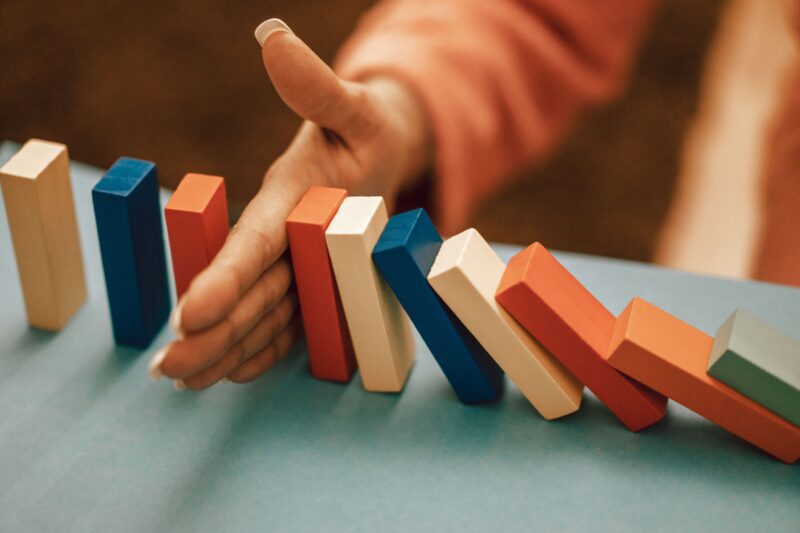 A hand placed between a row of falling coloured wooden blocks stops the chain reaction, leaving the remaining blocks standing, illustrating emergency egress as the final layer of protection when earlier safety layers have failed