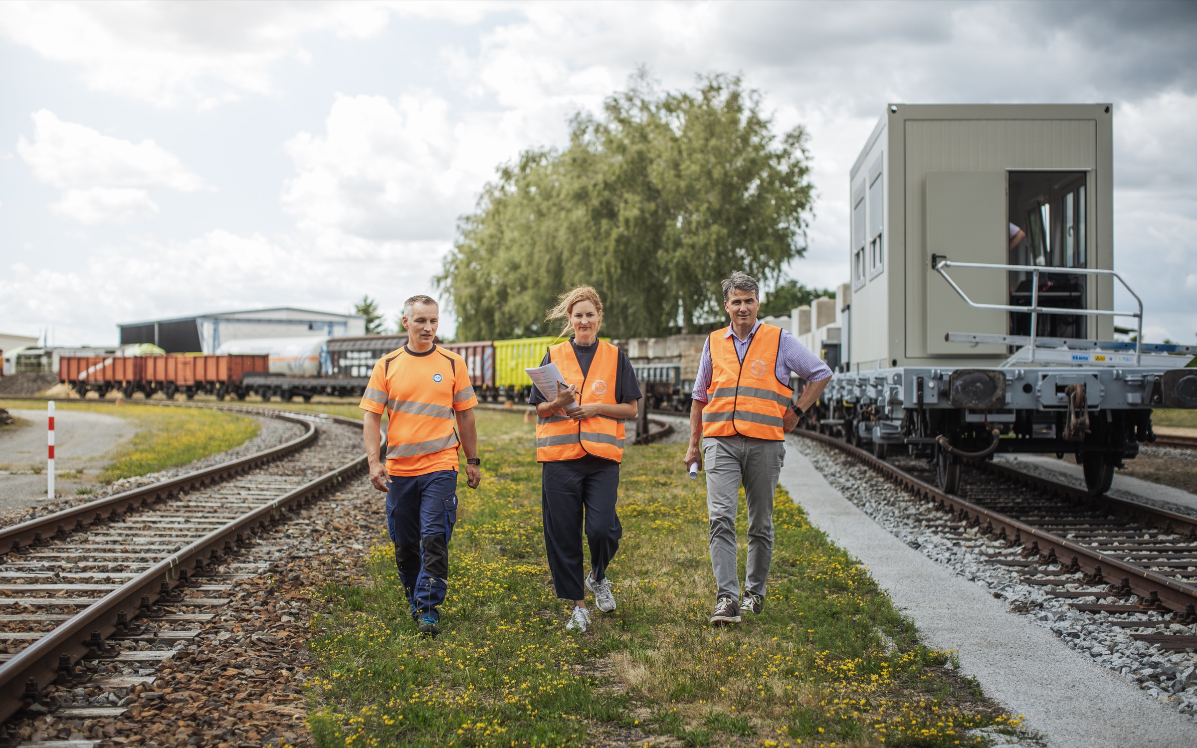 Three TÜV SÜD rail staff in high-visibility vests walking beside a freight wagon on a test track